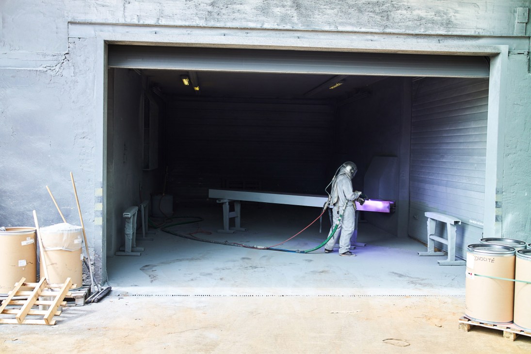Worker in protective gear spraying galvanized coating on metal street furniture in an industrial facility.