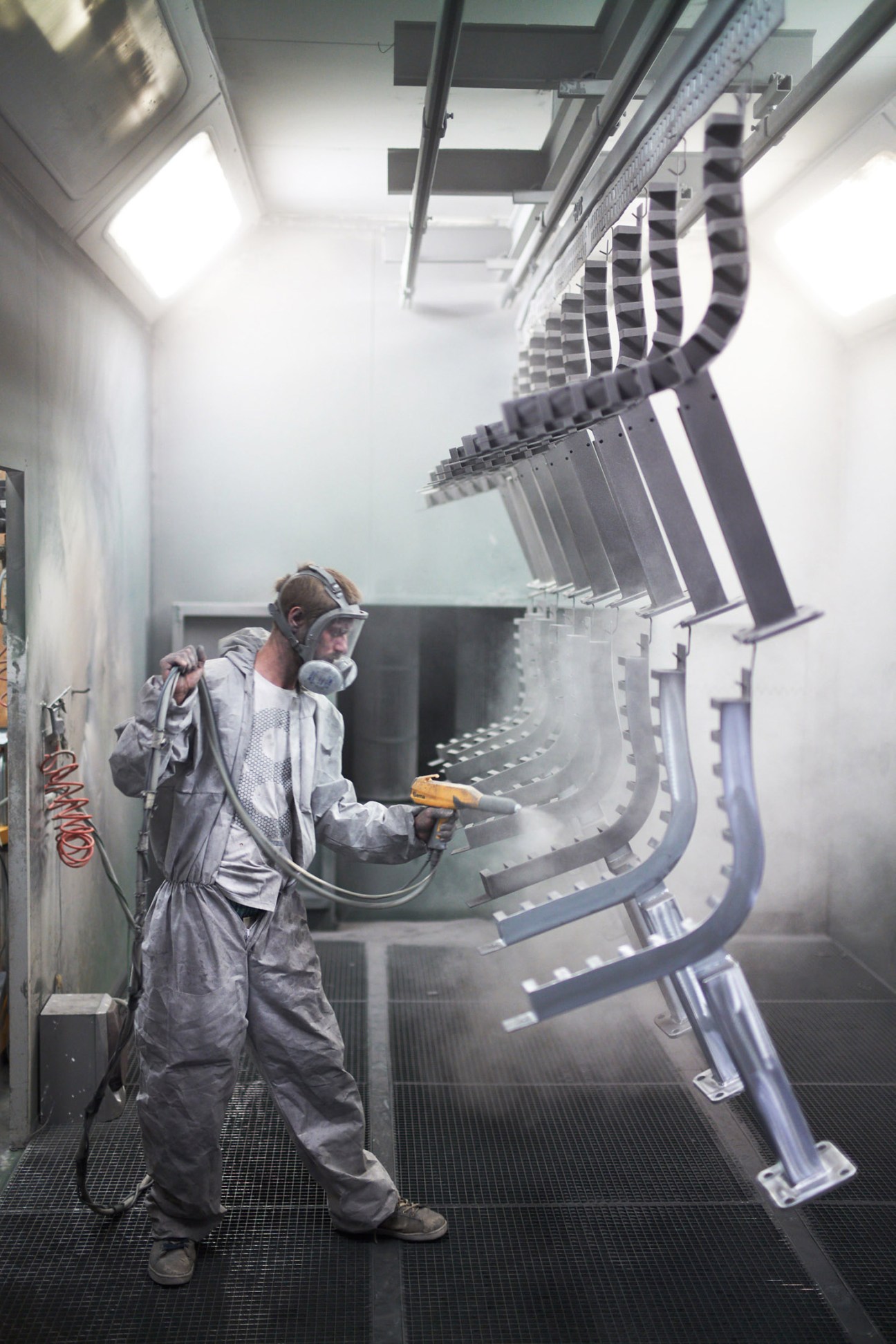 Worker in protective suit spray-coating steel street furniture components in industrial facility.