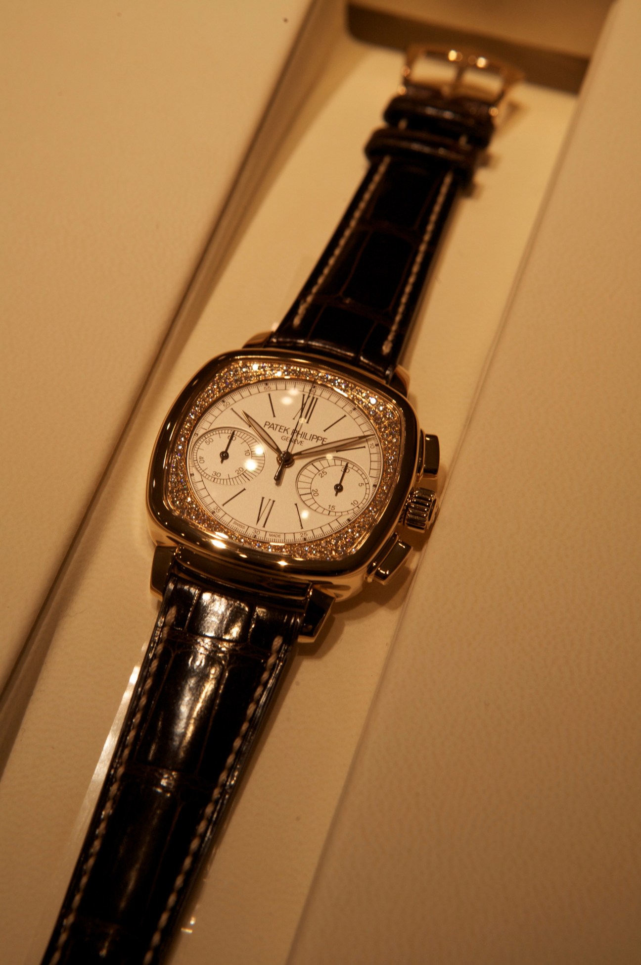 Bejewelled Patek Philippe timepiece with diamond-set bezel, cream dial, and black alligator strap on display.
