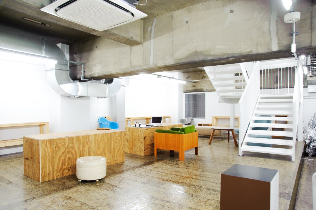 Raw industrial office space with concrete floors, exposed beams, wooden desks, and stairs.
