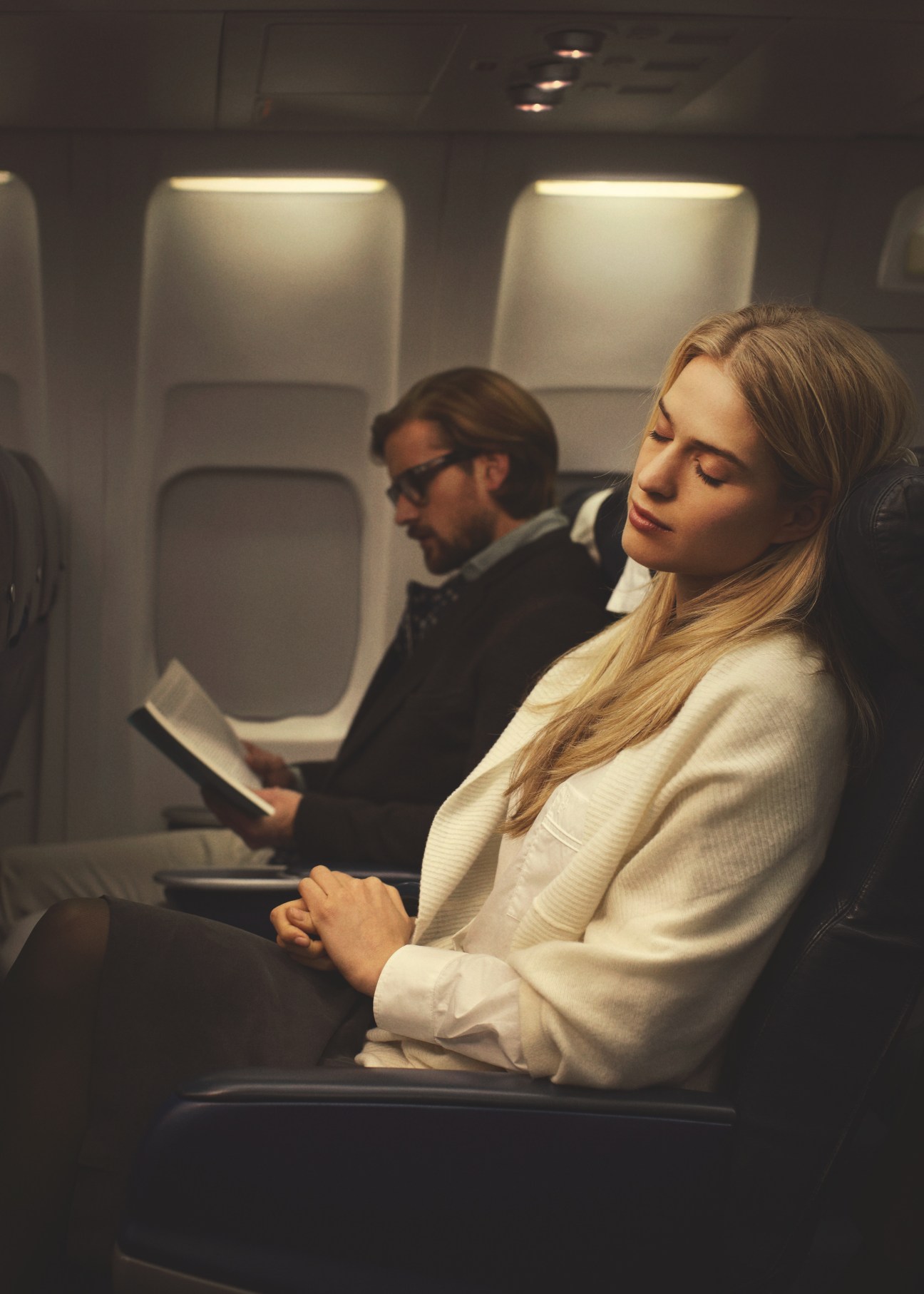 Woman in cream cape and Hermès shirt seated on airplane with passenger in background