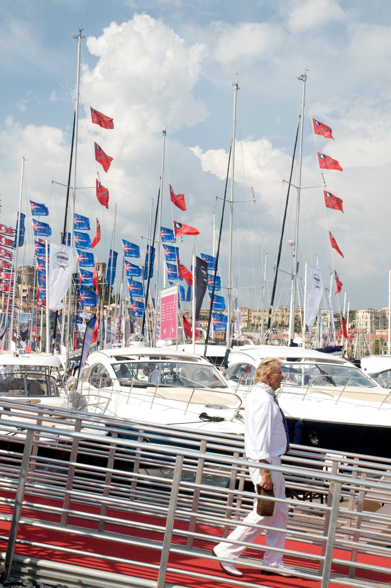 Person in white clothing walking on a dock past moored sailboats with red and blue flags.