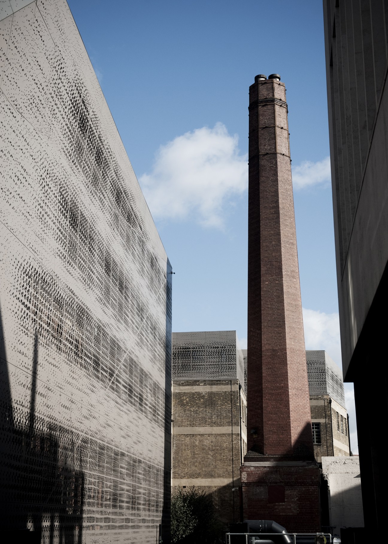 Modern perforated white building facade contrasts with historic red brick industrial chimney and traditional structures under blue sky.