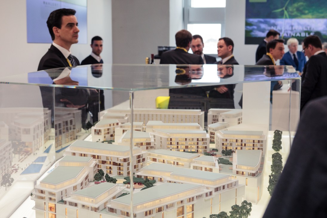 Businessmen in suits view an architectural model of residential buildings at the Stuttgart booth, with additional attendees visible in the background.