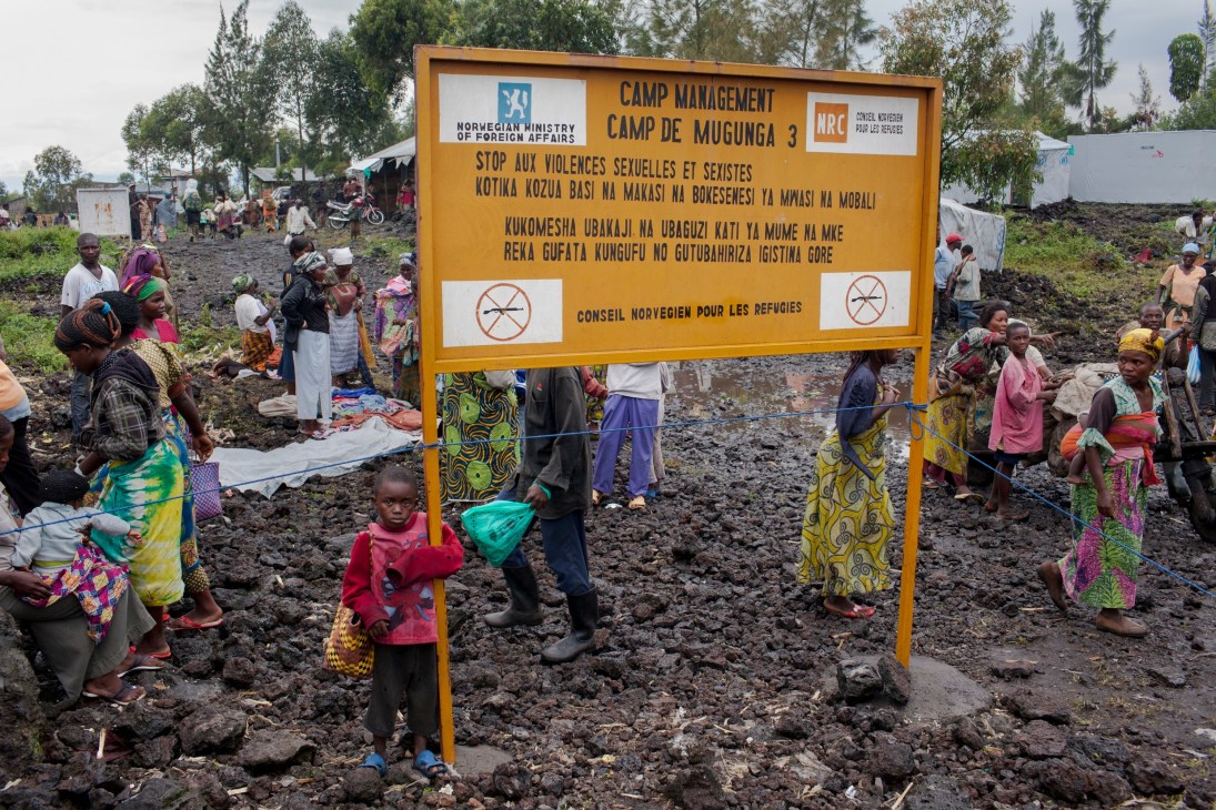 A yellow sign for Camp de Mugunga 3 refugee camp in the DRC surrounded by internally displaced people and makeshift shelters.