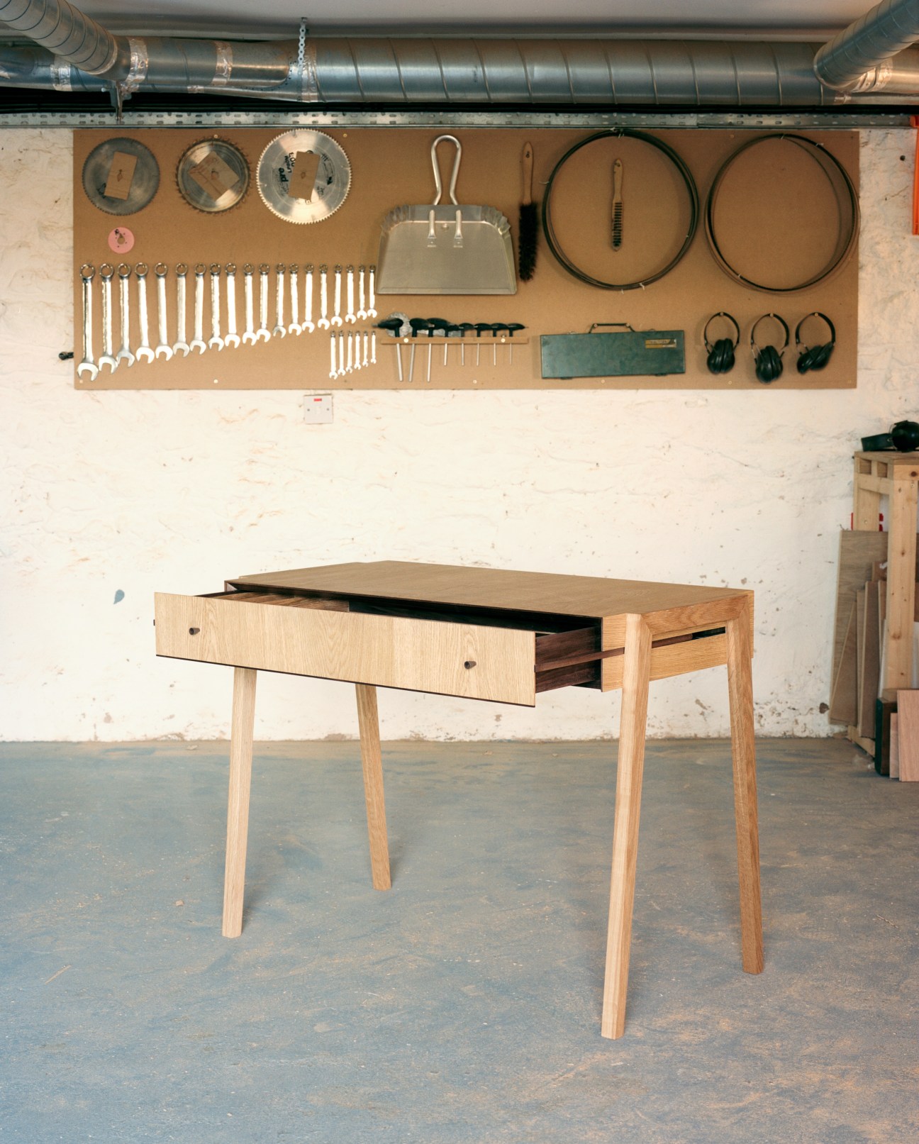 Handcrafted wooden desk with open drawer in a workshop with organized tools on pegboard wall.