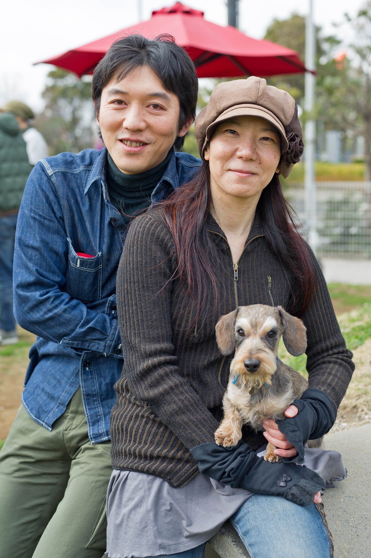 Mr and Mrs Takahashi pose outdoors with their small dog Raita under a red umbrella.