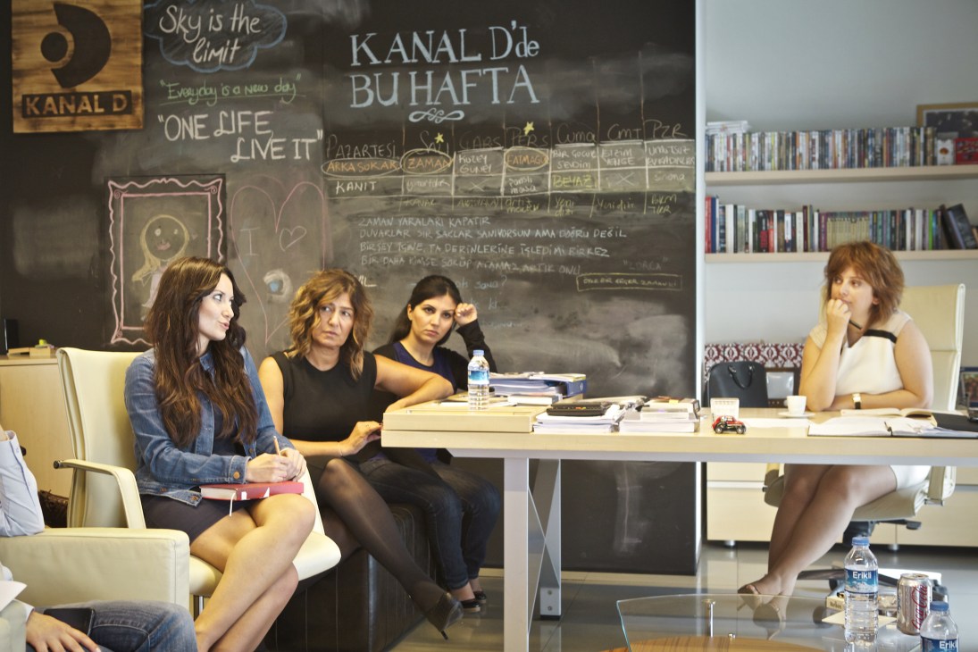 Four women in a Kanal D office discuss a proposal beside a chalkboard with Turkish text and series information.