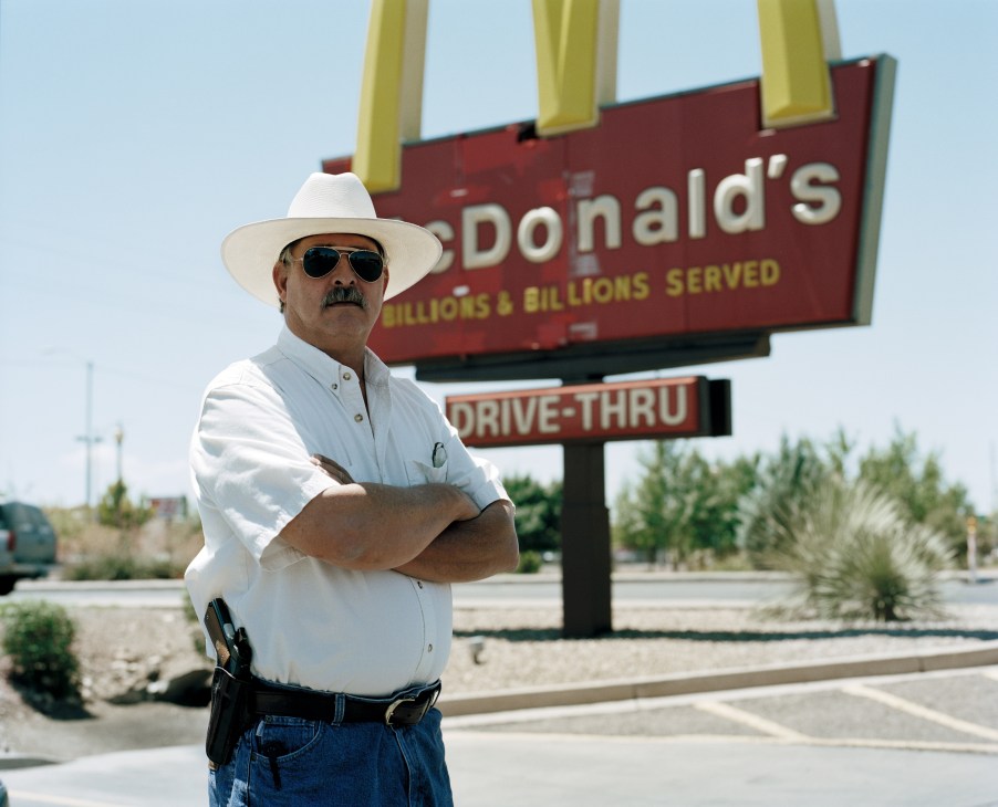 Man in white cowboy hat and sunglasses standing with arms crossed in front of McDonald's sign, wearing holstered gun.