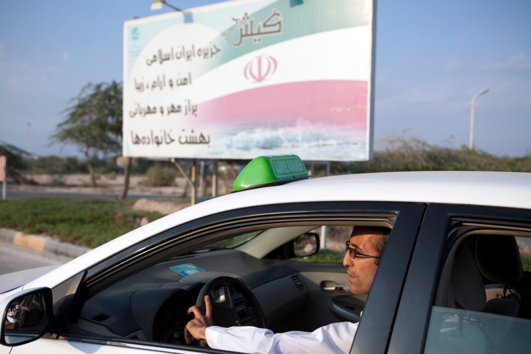 Taxi driver in a white car with green roof light in front of an Iranian government sign.