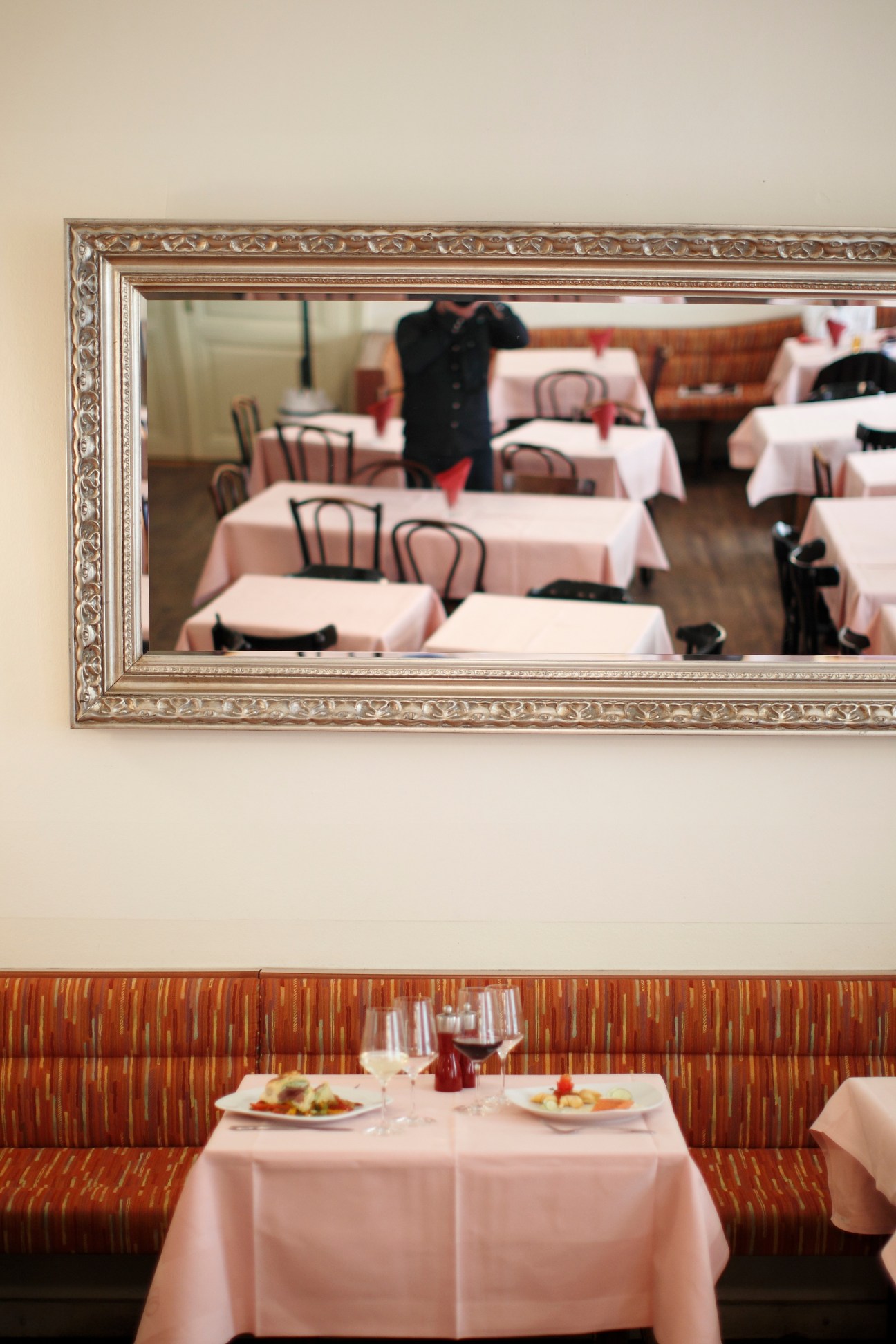 Restaurant interior with pink tablecloths, red striped banquette seating, and an ornate framed mirror on the wall reflecting the dining room.