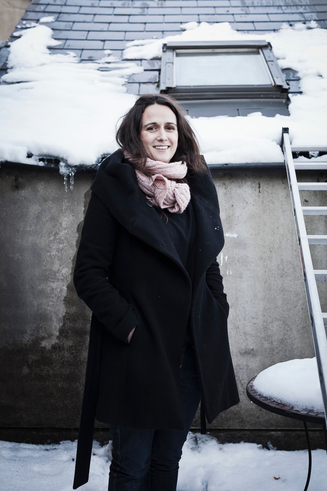 Miranda Driscoll stands smiling in front of a snow-covered roof with a skylight, wearing a black coat and pink scarf.
