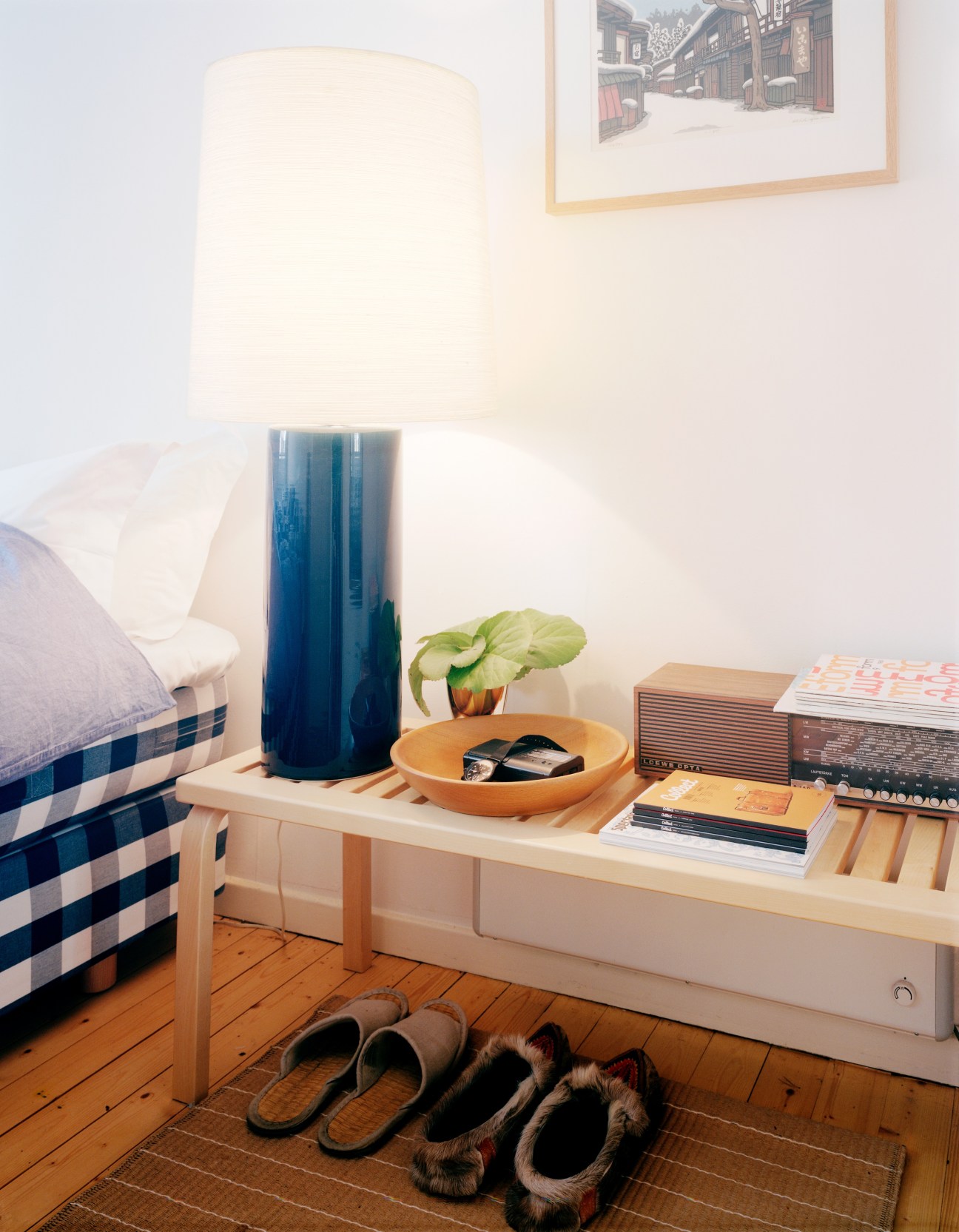 Bedroom with Hästens bed, Artek table, ceramic lamp with blue base, wooden shelf with magazines, and elk boots on floor.