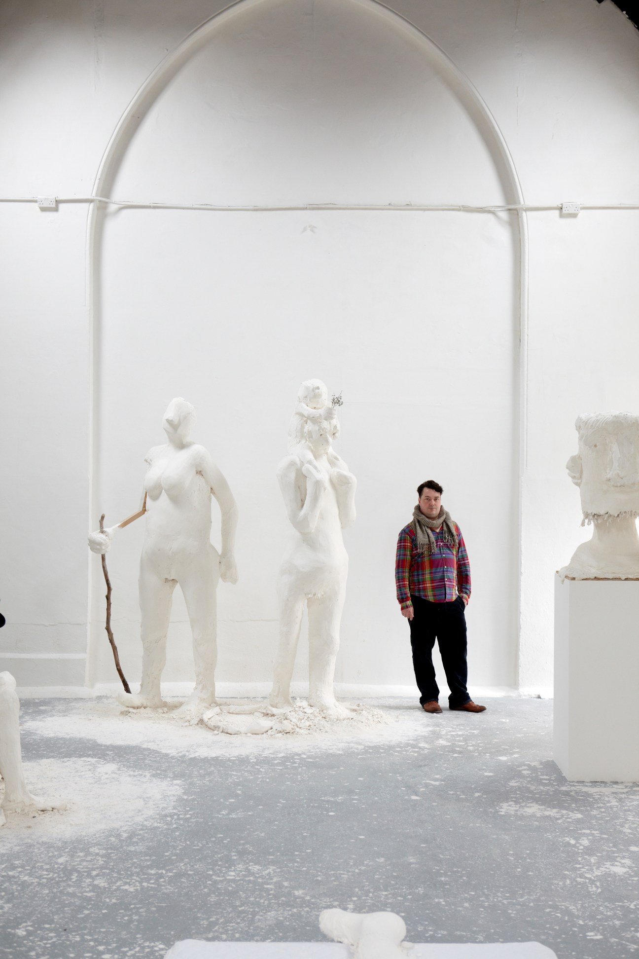 Man in plaid shirt stands among white plaster sculptures in an arched gallery space.