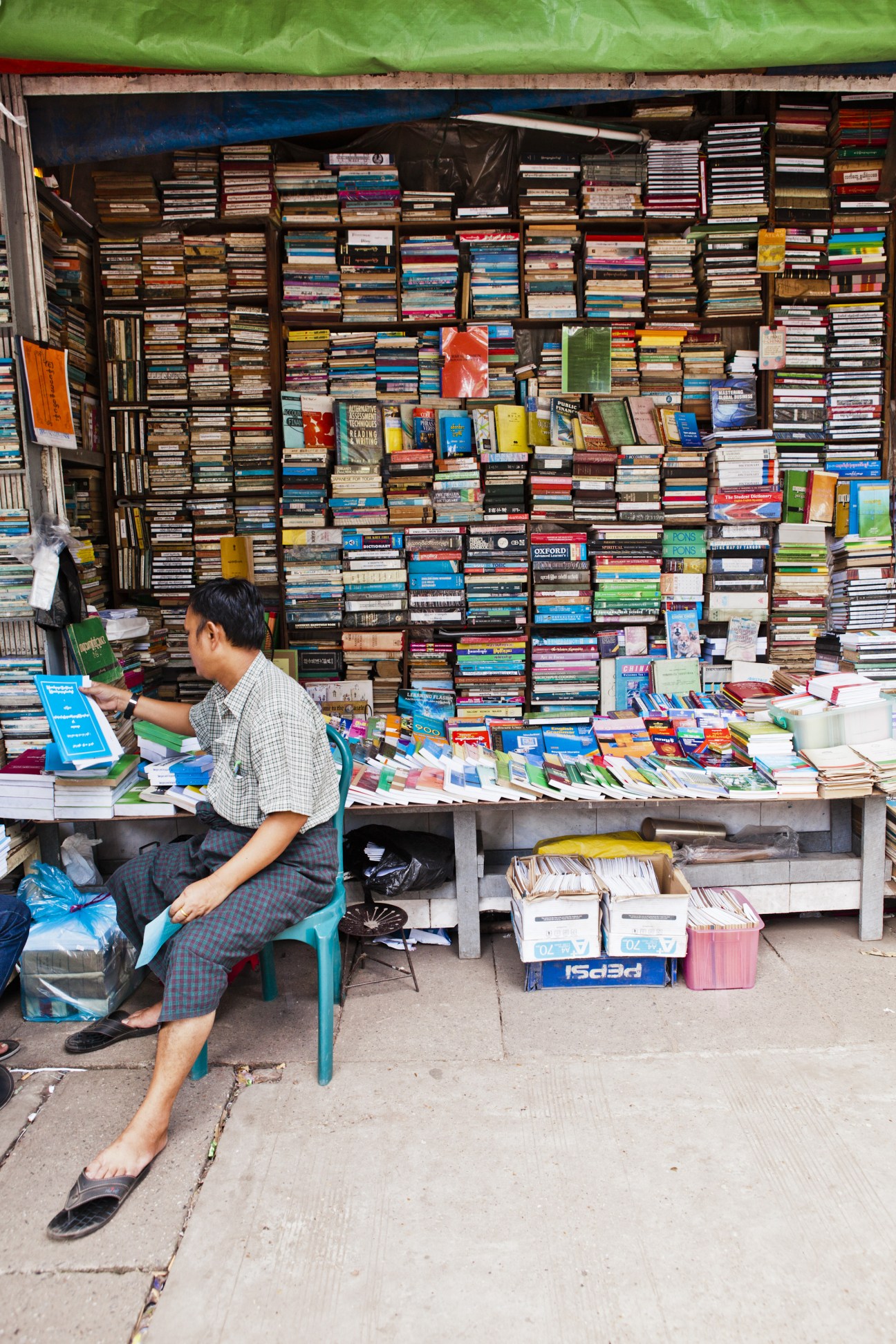 A man sits at a street bookstall in Rangoon with hundreds of books stacked on shelves behind him.