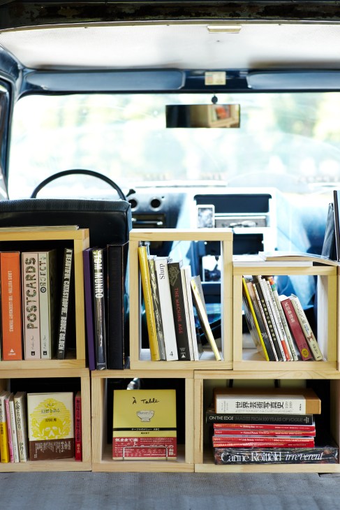 Interior of a vintage Chevy Van displaying books and magazines in wooden shelving units, viewed from the driver's seat.