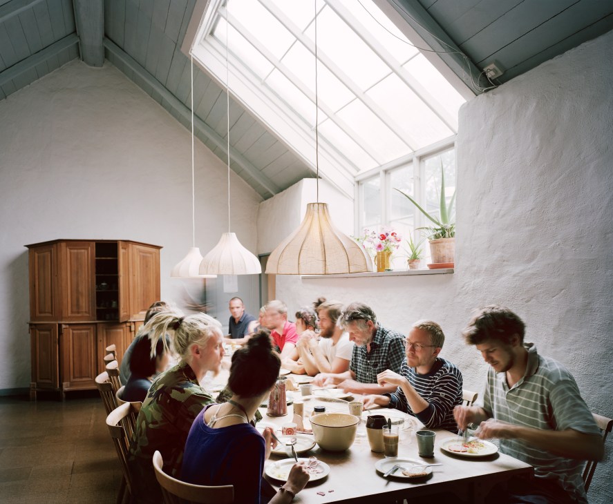 Students dining together at a long table in a bright, minimalist room with pendant lights and wooden furniture.