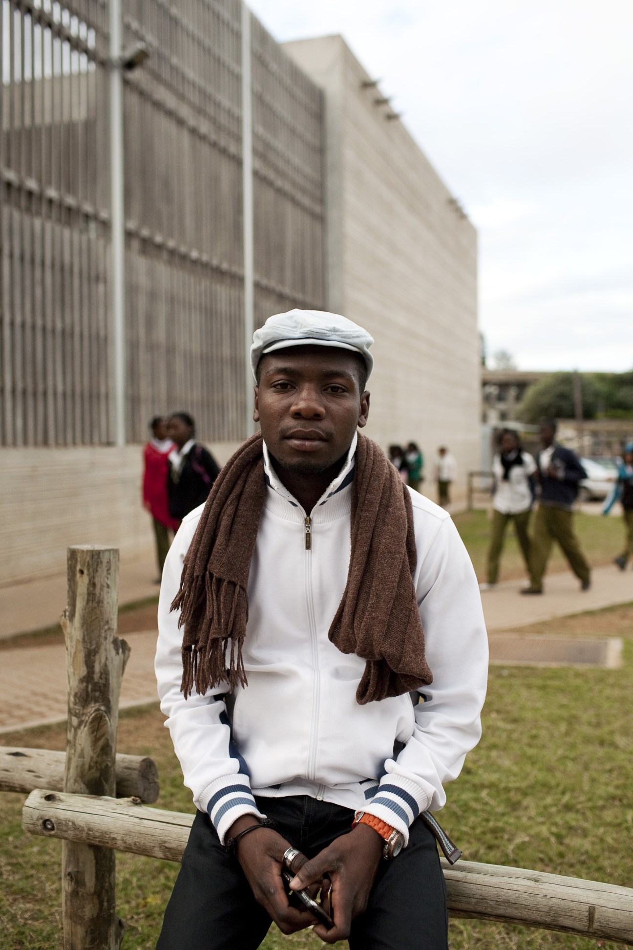 A man in a white zip-up hoodie and flat cap sits on a wooden bench outside a modern concrete building.