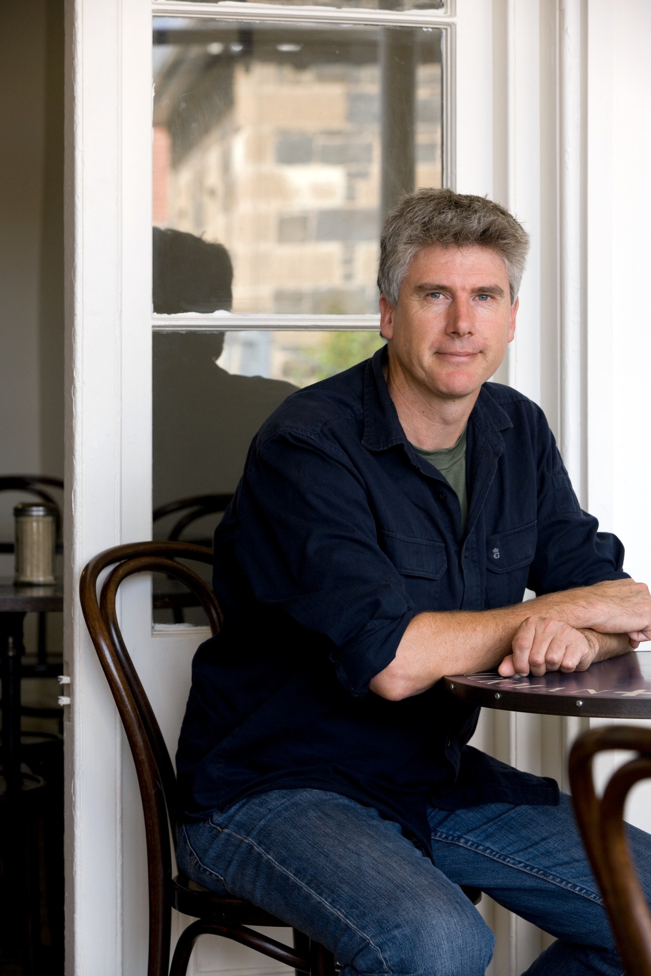 Matthew Evans seated at a table by a window, wearing a dark navy shirt over a green t-shirt.
