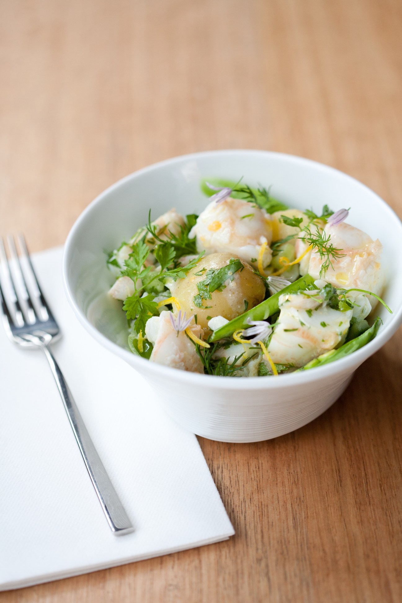 A white bowl of seafood dish with herbs and edible flowers on a wooden table with fork.