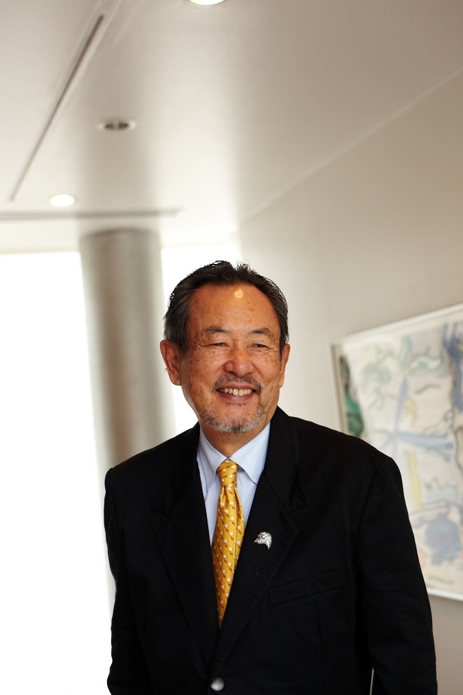 Toshio Hara, founder of the Hara Museum of Contemporary Art in Tokyo, standing in a modern gallery space wearing a dark suit and gold tie.