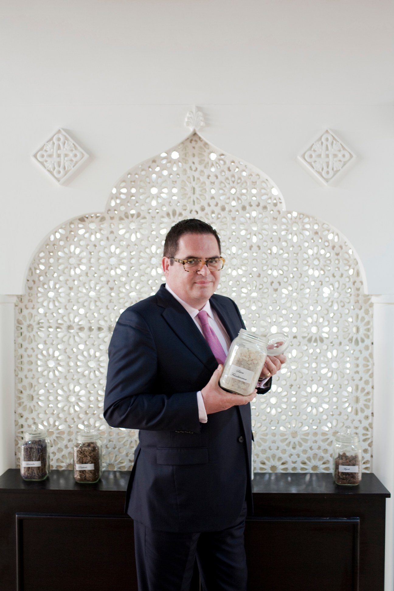 David Crickmore, CEO of Amouage, holds a jar of perfume in front of an ornate white geometric backdrop.
