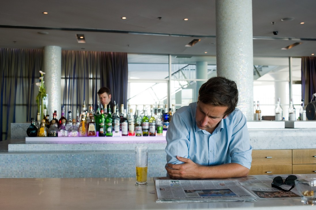 Man in blue shirt reading newspaper at bar counter with drink and bottles visible behind