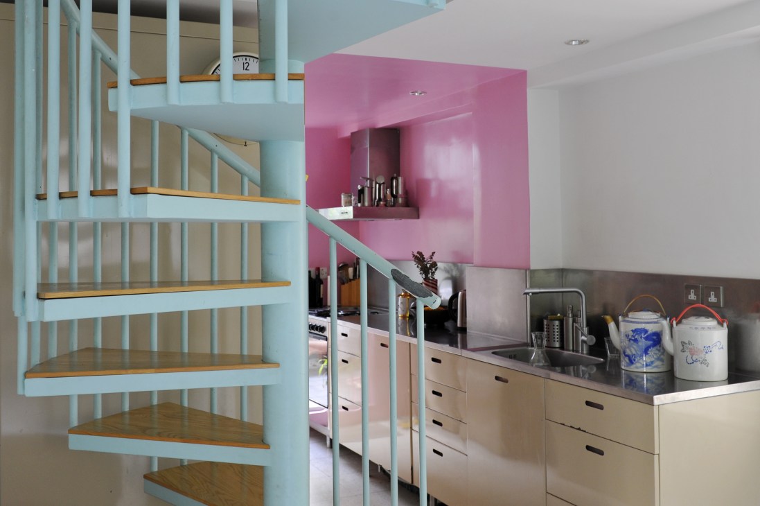 Modern kitchen with light blue spiral staircase, pink accent wall, wooden countertops and storage drawers.
