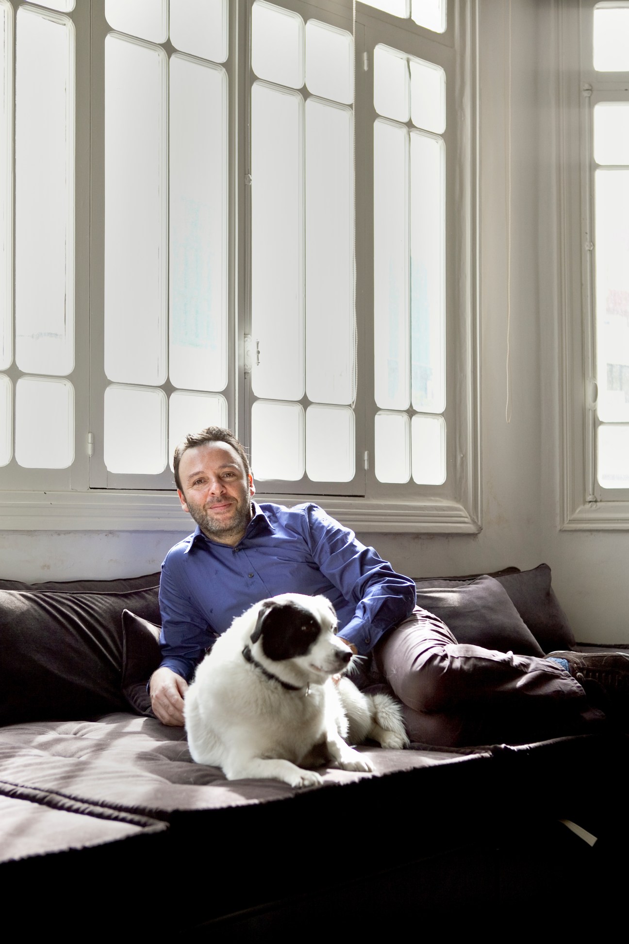 Man in blue shirt sitting on dark leather sofa with white and black dog, large multi-paned windows behind.