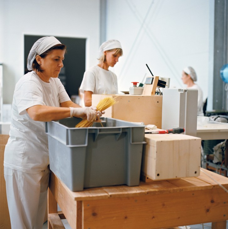 Factory worker in white uniform and hairnet sorts dried spaghetti pasta into containers at a production facility.