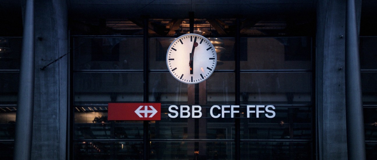 Illuminated analog clock above SBB CFF FFS railway signage on dark building facade.