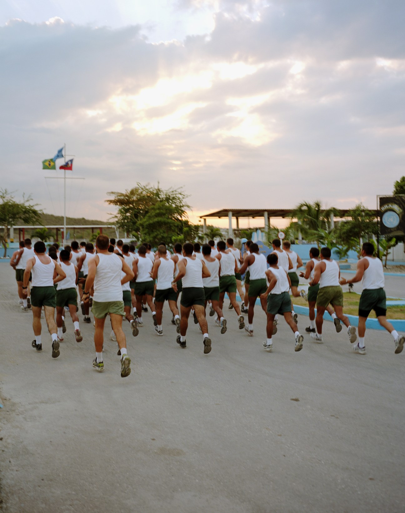 Brazilian soldiers in white tank tops and olive shorts run together around a base courtyard under flags.