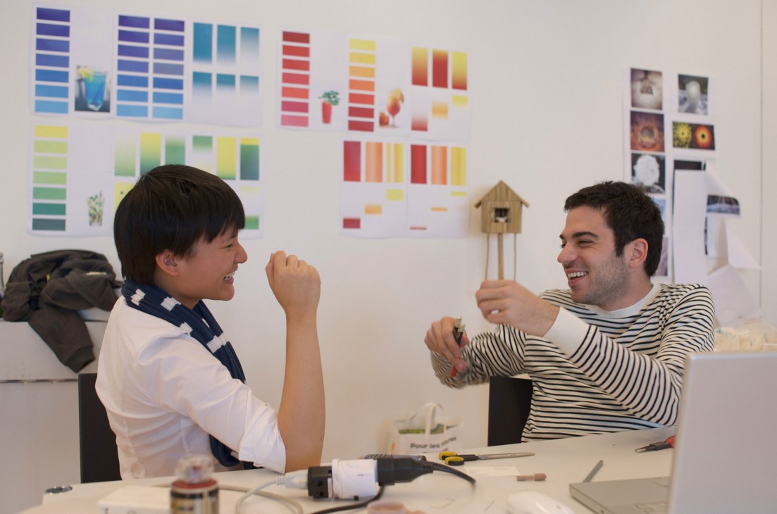 Two ECAL students laugh together at a desk with design work and colorful swatches displayed on the wall behind them.