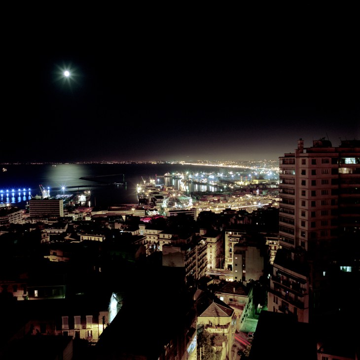 Nighttime aerial view of Algiers with illuminated harbor, ships, and suburban construction visible under moonlight.
