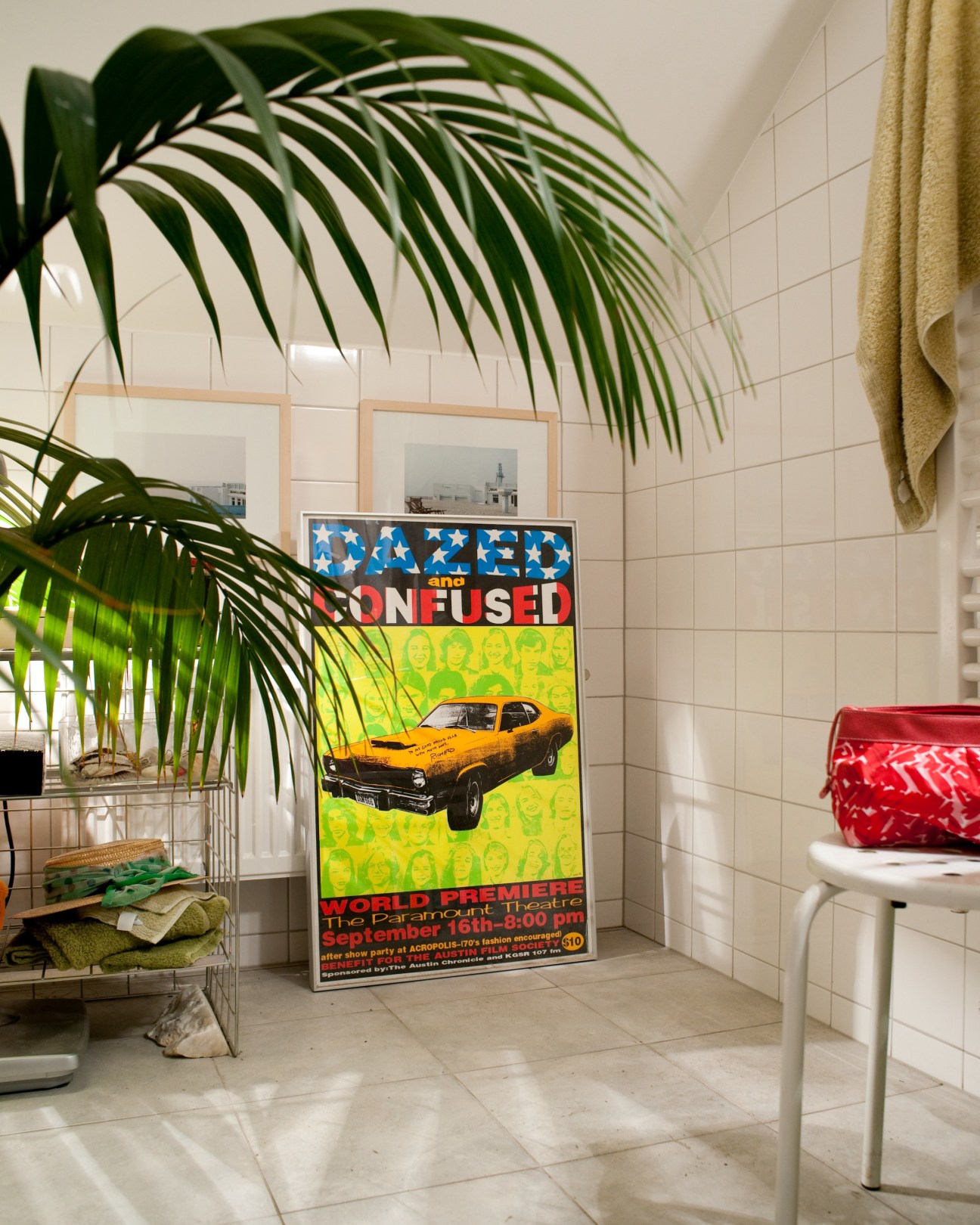 A colorful film poster for 'Dazed and Confused' displayed in a bathroom with white tiles, potted plant, and towels.