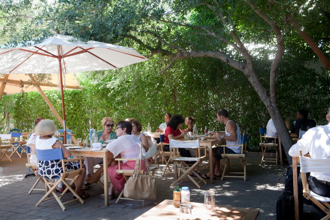 Diners seated at wooden tables under umbrellas and shade trees at an outdoor restaurant patio.