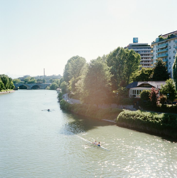 Rowers in single sculls on the river Po with bridges, trees, and Turin buildings in background.