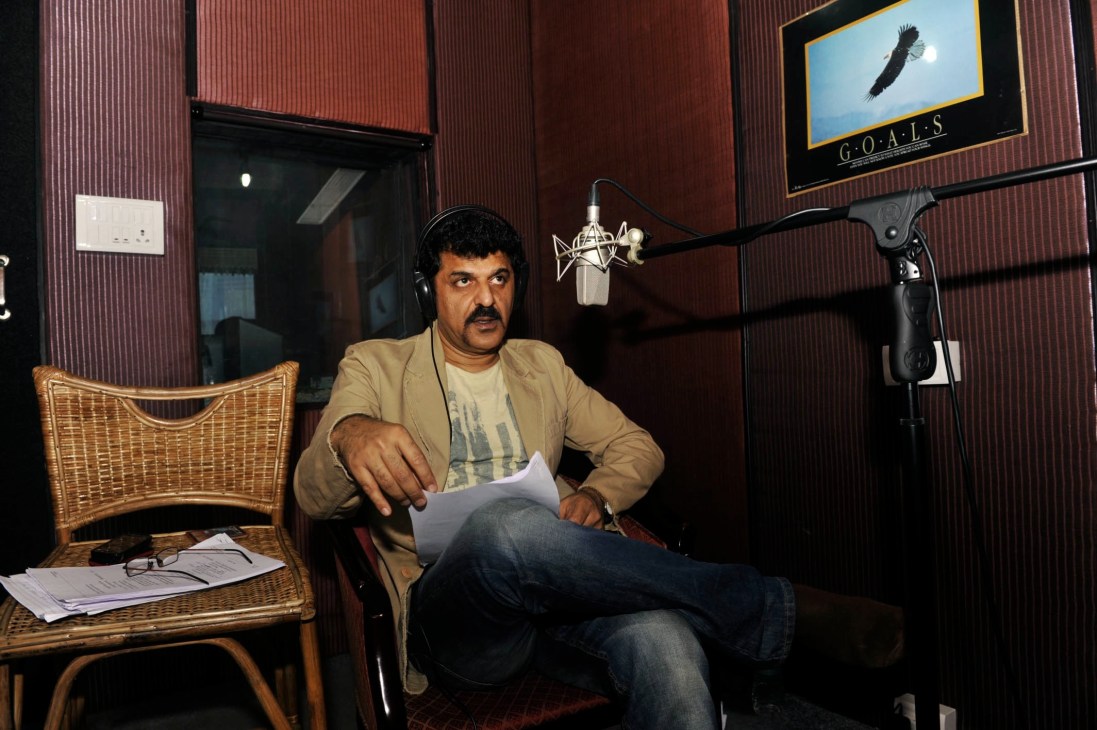 Rajesh Khattar sits in a recording studio wearing headphones, holding a script while positioned at a professional microphone.