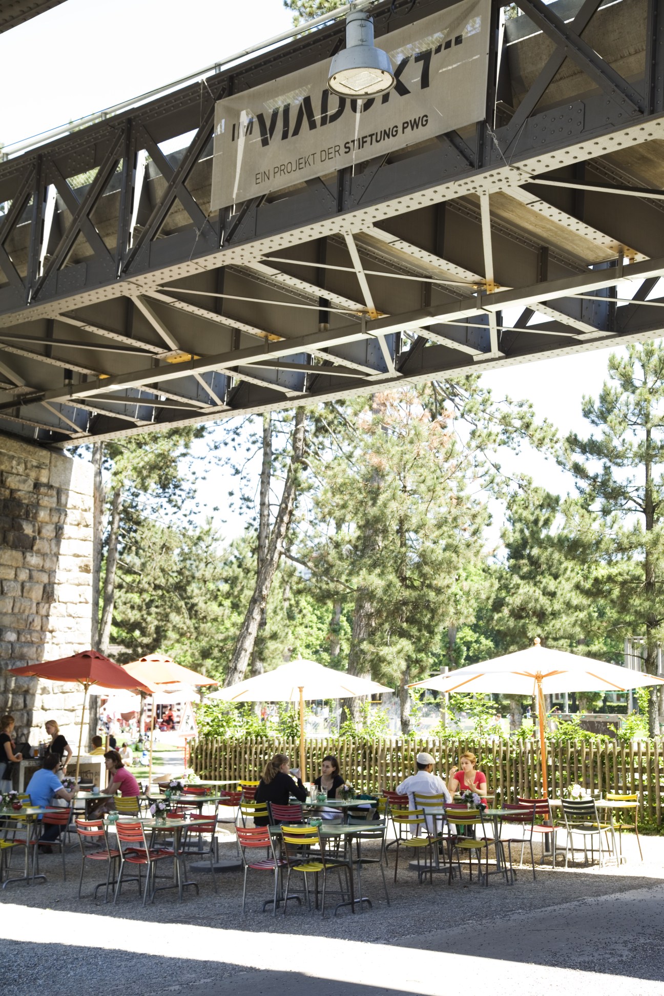 Outdoor café seating under industrial metal bridge structure with umbrellas and trees.