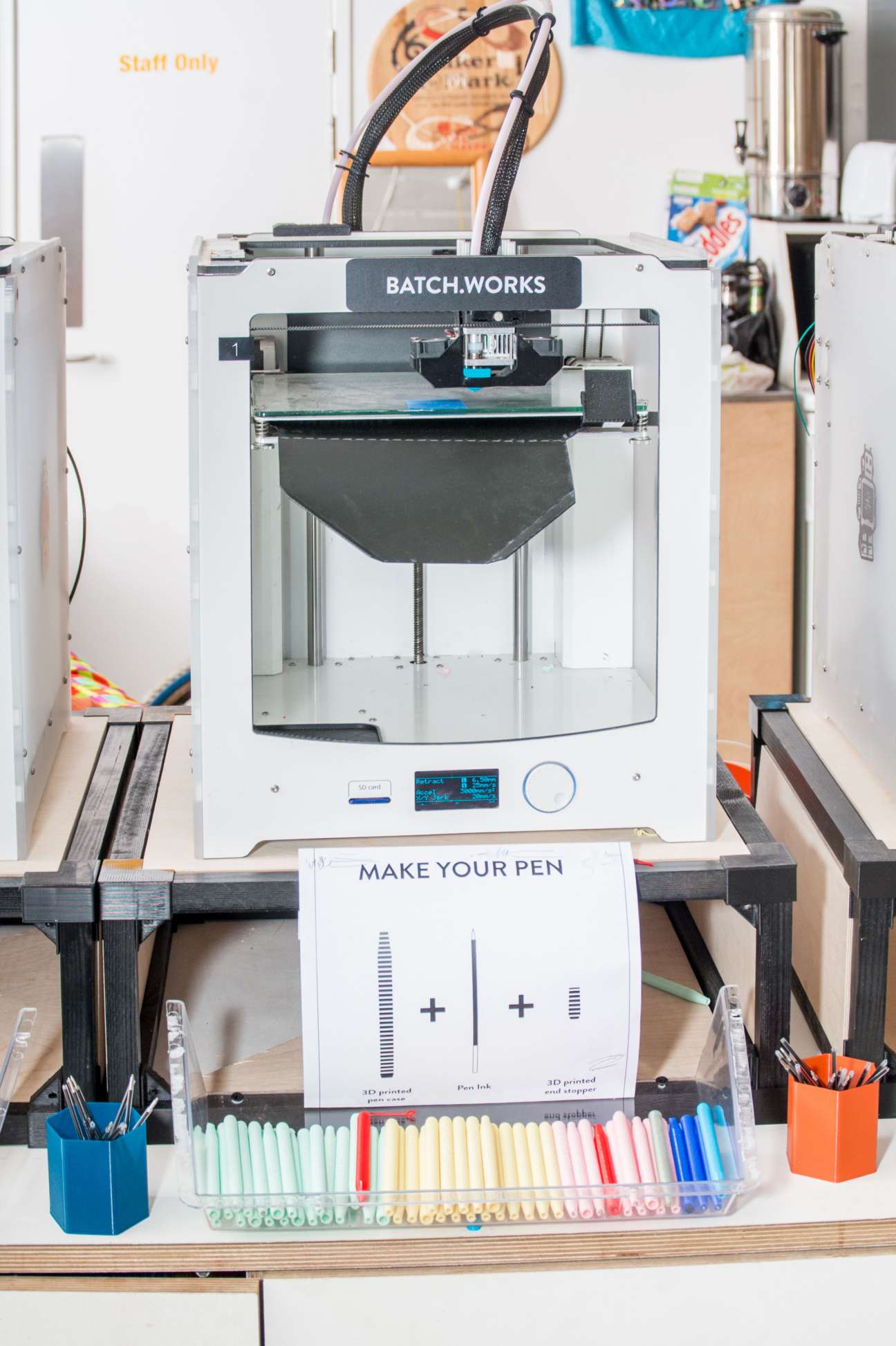 A white BatchWorks 3D printer on a desk with colored markers and a 'Make Your Pen' instruction card below it.