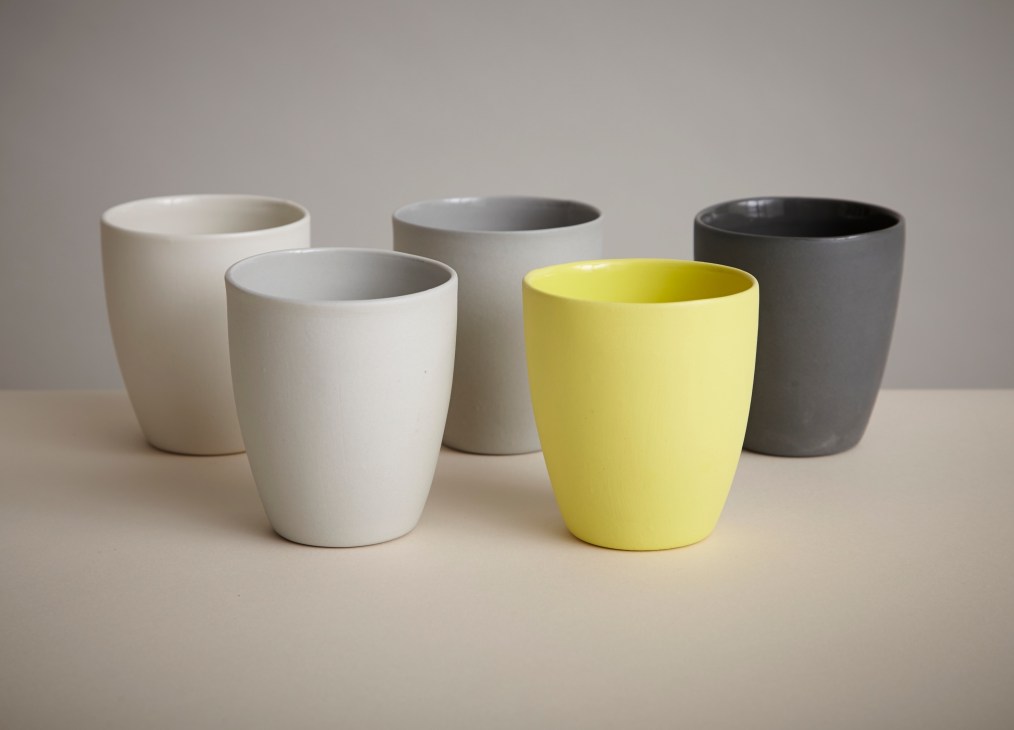 Five handmade ceramic cups in white, cream, yellow and gray displayed on a neutral surface.