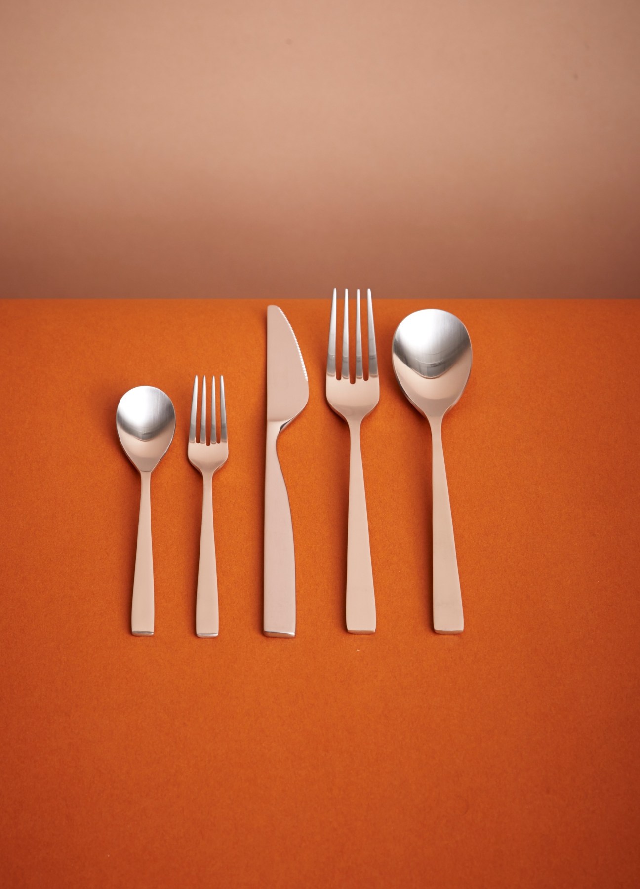 Five pieces of stainless steel cutlery arranged on an orange background against a gradient wall.