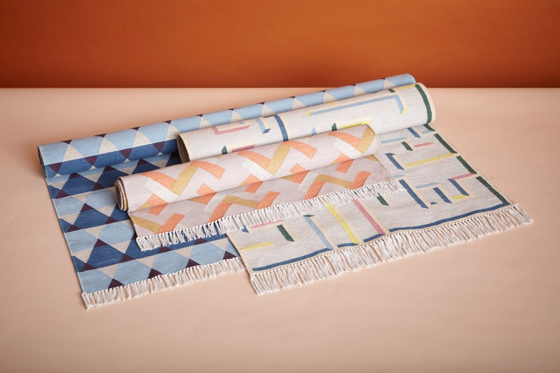 Three hand-woven cotton dhurries with geometric patterns displayed on a beige surface against an orange background.