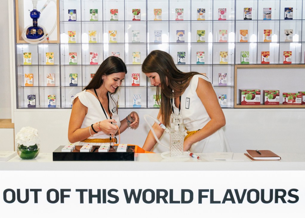 Two women in white uniforms examining tea or spice products at a duty-free retail display with colorful packages
