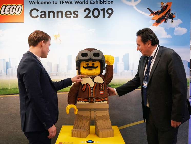 Two businessmen in suits shake hands over a large LEGO figure display at the TFWA World Exhibition in Cannes 2019