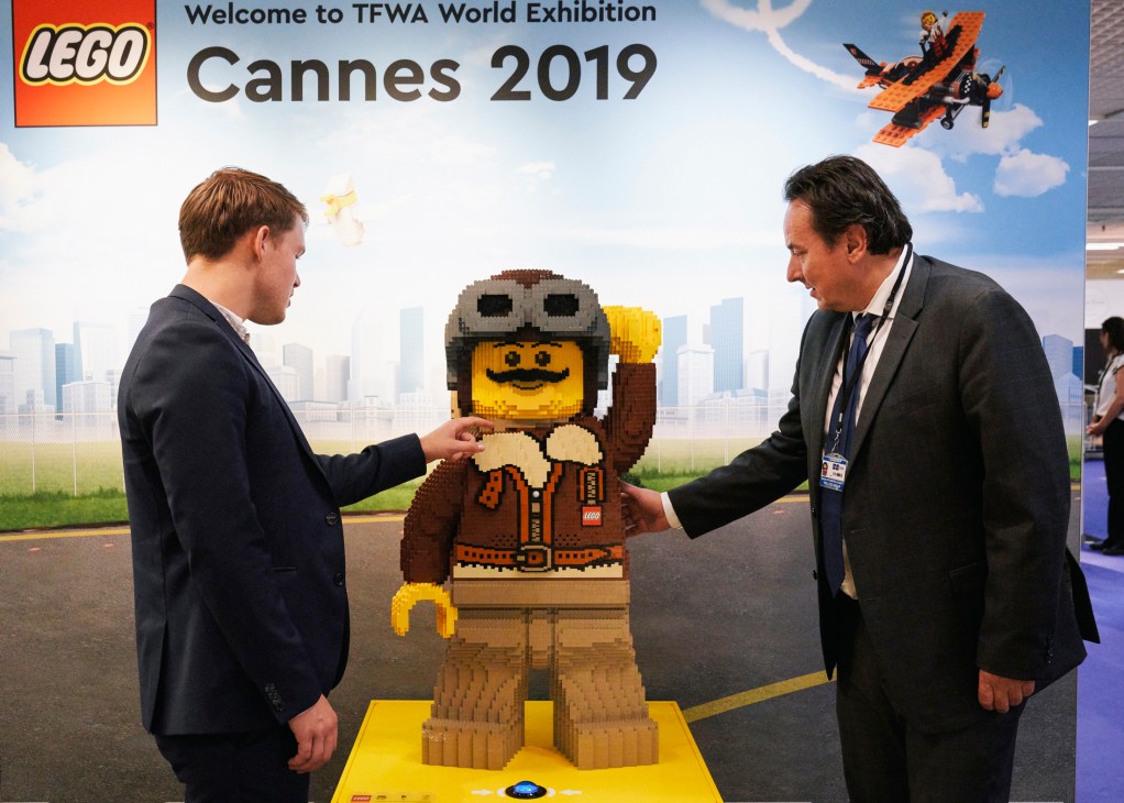 Two businessmen in suits shake hands over a large LEGO figure display at the TFWA World Exhibition in Cannes 2019