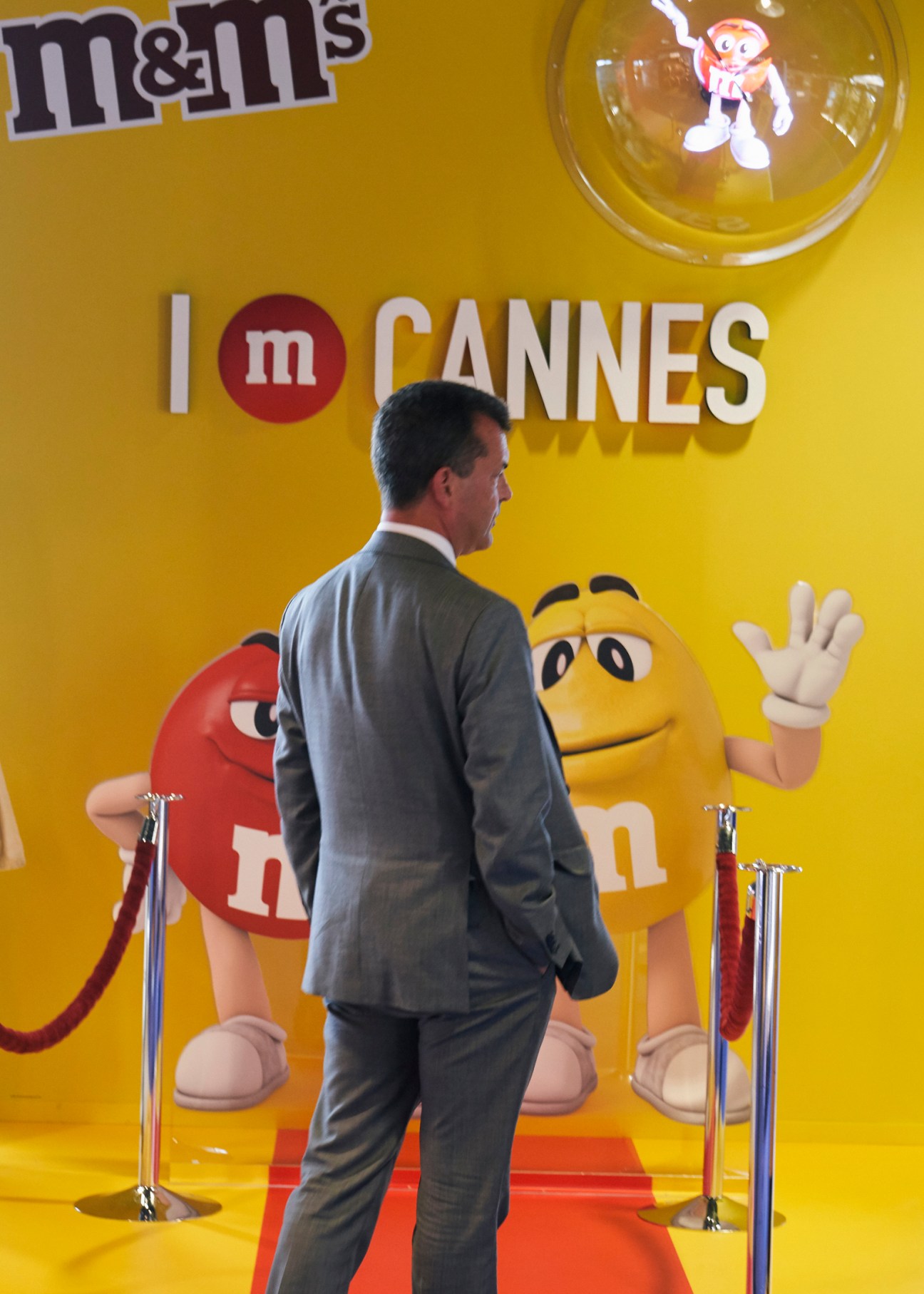 Man in grey suit walking on red carpet past large yellow M&M's branded display at trade show