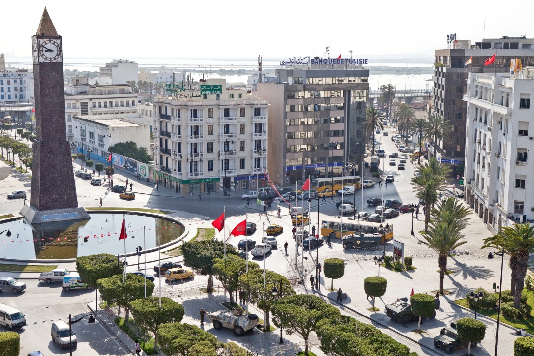 Aerial view of Avenue Bourguiba in Tunis showing a clock tower, colonial buildings, palm trees, red flags, and people gathering in the main square.