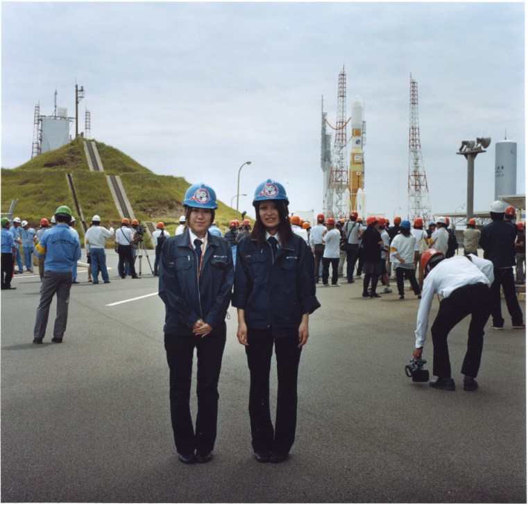 Two female JAXA guides in blue hard hats and uniforms stand at a launch pad surrounded by staff and media.