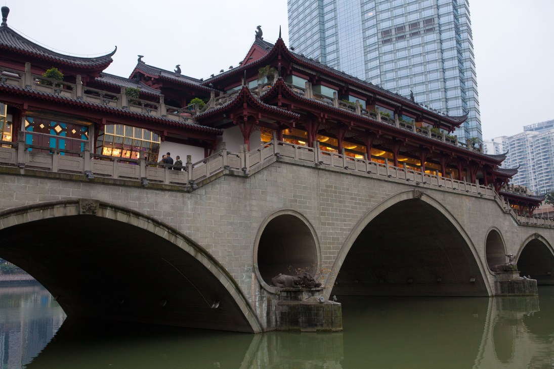 Traditional Chinese temple with red-tiled roofs spanning a stone bridge over water, with modern high-rise buildings visible behind.